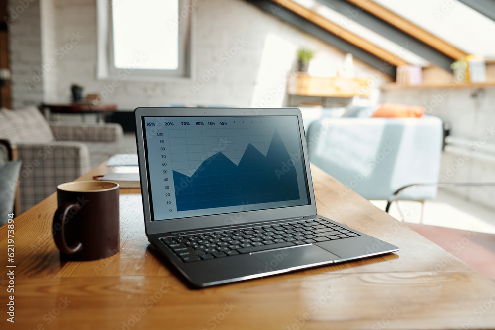 Laptop with blue financial graph on screen and brown mug with tea or coffee standing on table against part of spacious coworking area Laptop with blue financial graph on screen and brown mug with tea or coffee standing on table against part of spacious coworking area