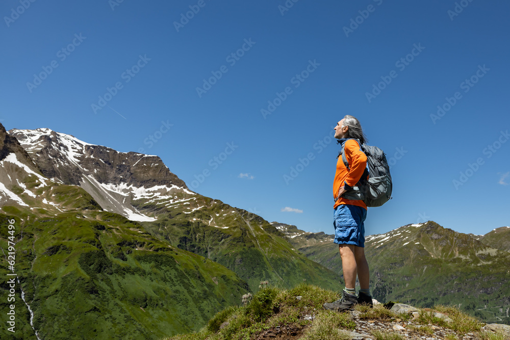 Adult male hiker with backpack standing at a height and looking at a mountain landscape with snow-capped peaks, Austria