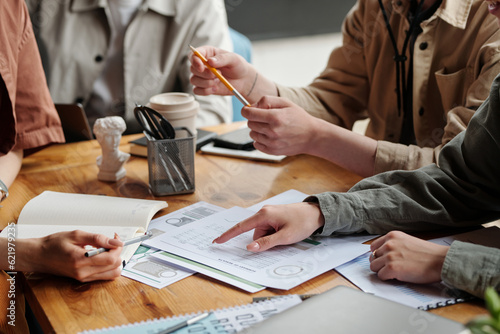 Hand of young female economist pointing at financial document and explaining data to colleagues sitting in front of her at working meeting