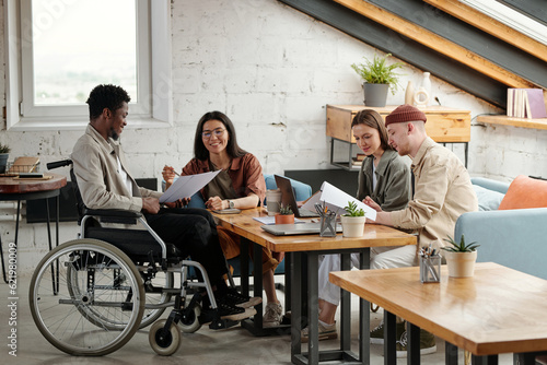 Two pairs of employees working in small groups by workplace while female economist pointing at document held by male colleague in wheelchair photo