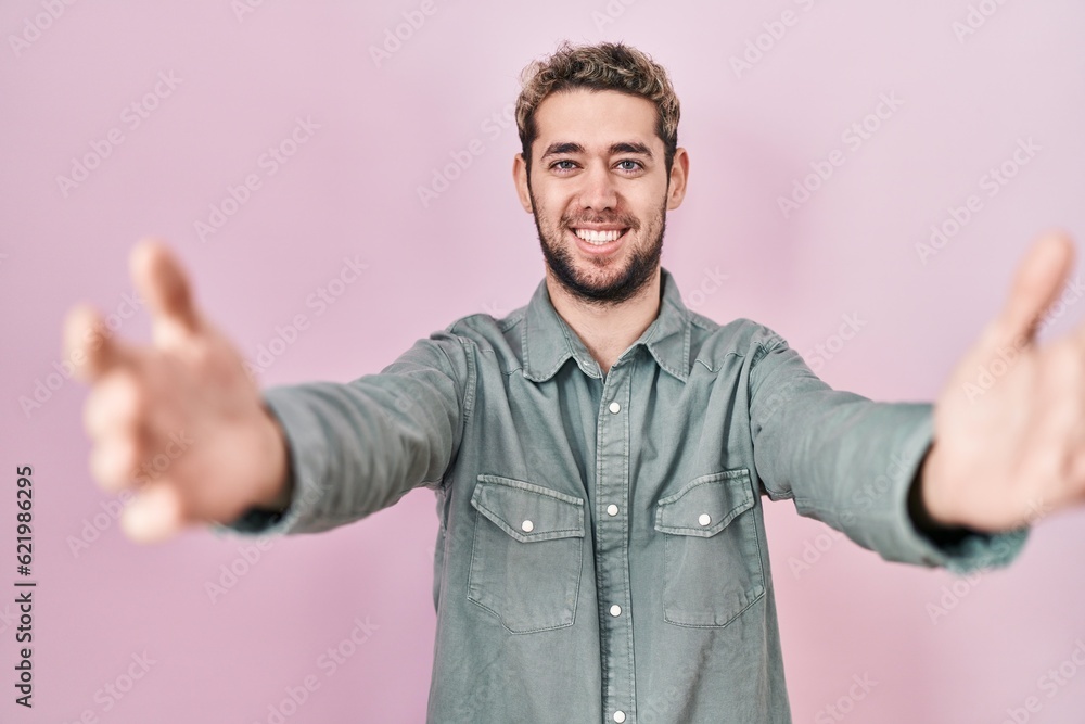 Hispanic man with beard standing over pink background looking at the camera smiling with open arms for hug. cheerful expression embracing happiness. Hispanic man with beard standing over pink background looking at the camera smiling with open arms for hug. cheerful expression embracing happiness.