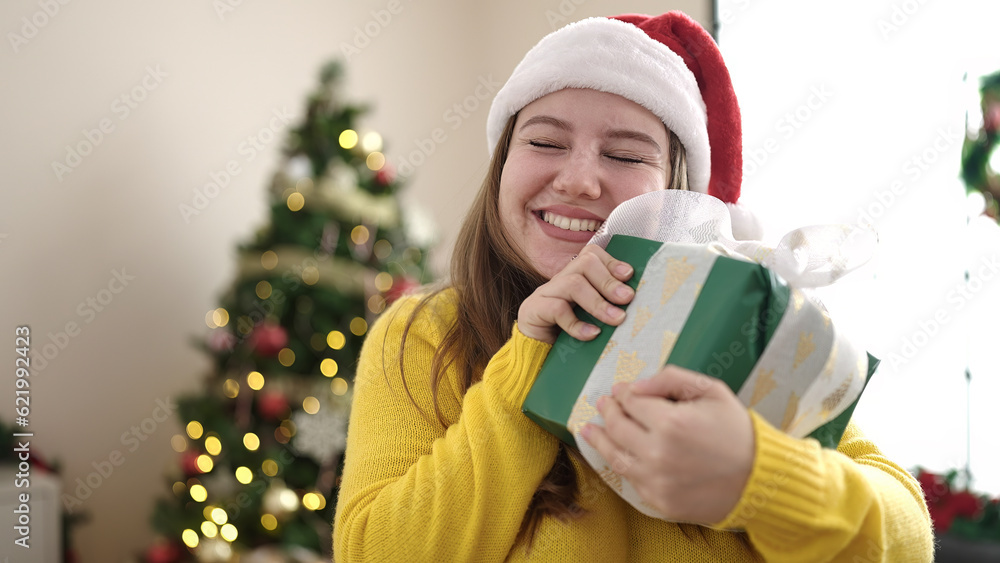 Young blonde woman holding gift standing by christmas tree at home Young blonde woman holding gift standing by christmas tree at home