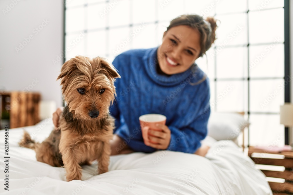 Young beautiful hispanic woman drinking cup of coffee sitting on bed with dog at bedroom Young beautiful hispanic woman drinking cup of coffee sitting on bed with dog at bedroom