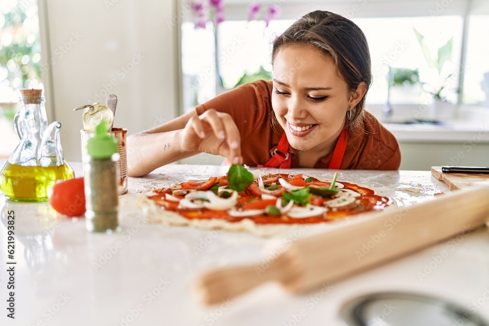 Young beautiful hispanic woman smiling confident putting basil on pizza at the kitchen Young beautiful hispanic woman smiling confident putting basil on pizza at the kitchen