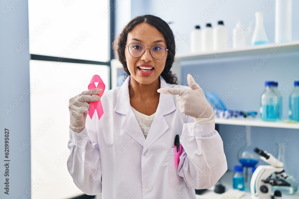 Young hispanic doctor woman working at scientist laboratory holding pink ribbon smiling happy pointing with hand and finger Young hispanic doctor woman working at scientist laboratory holding pink ribbon smiling happy pointing with hand and finger