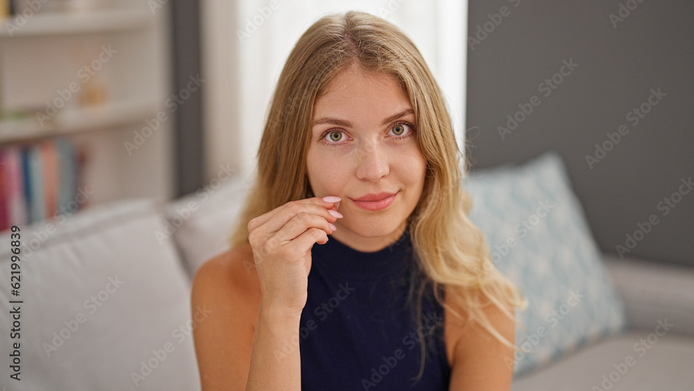 Young blonde woman sitting on sofa doing closing mouth gesture at home Young blonde woman sitting on sofa doing closing mouth gesture at home