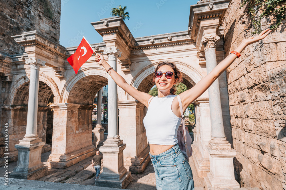 Happy traveller girl with turkish flag in hand and famous gate or Hadrian arch in Antalya. National holiday and must-visit tourist and sightseeing sites in Turkey