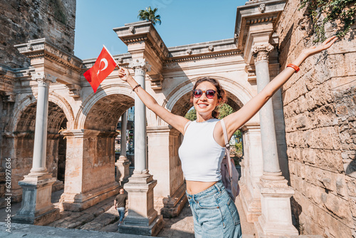 Happy traveller girl with turkish flag in hand and famous gate or Hadrian arch in Antalya. National holiday and must-visit tourist and sightseeing sites in Turkey