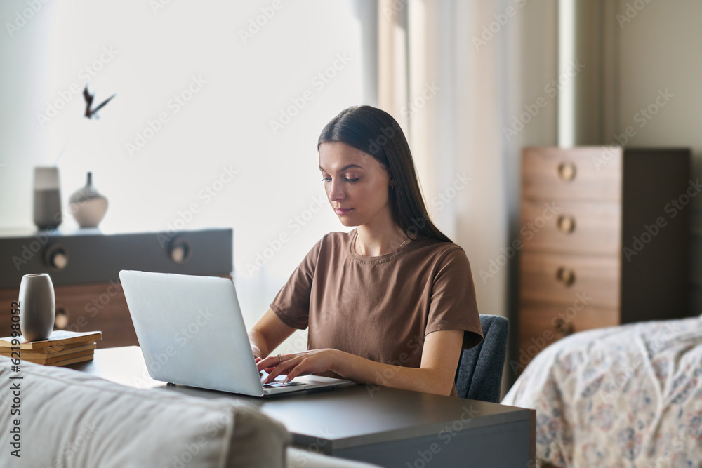 Young serious brunette businesswoman in t-shirt networking by desk in front of laptop while sitting against bed and interior of bedroom Young serious brunette businesswoman in t-shirt networking by desk in front of laptop while sitting against bed and interior of bedroom