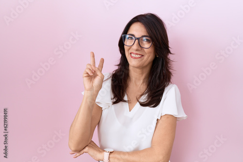 Middle age hispanic woman wearing casual white t shirt and glasses smiling with happy face winking at the camera doing victory sign with fingers. number two. © Krakenimages.com
