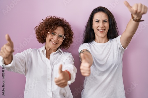 Hispanic mother and daughter wearing casual white t shirt over pink background approving doing positive gesture with hand, thumbs up smiling and happy for success. winner gesture.