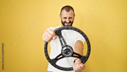 Young hispanic man smiling confident using steering wheel as a driver over isolated yellow background