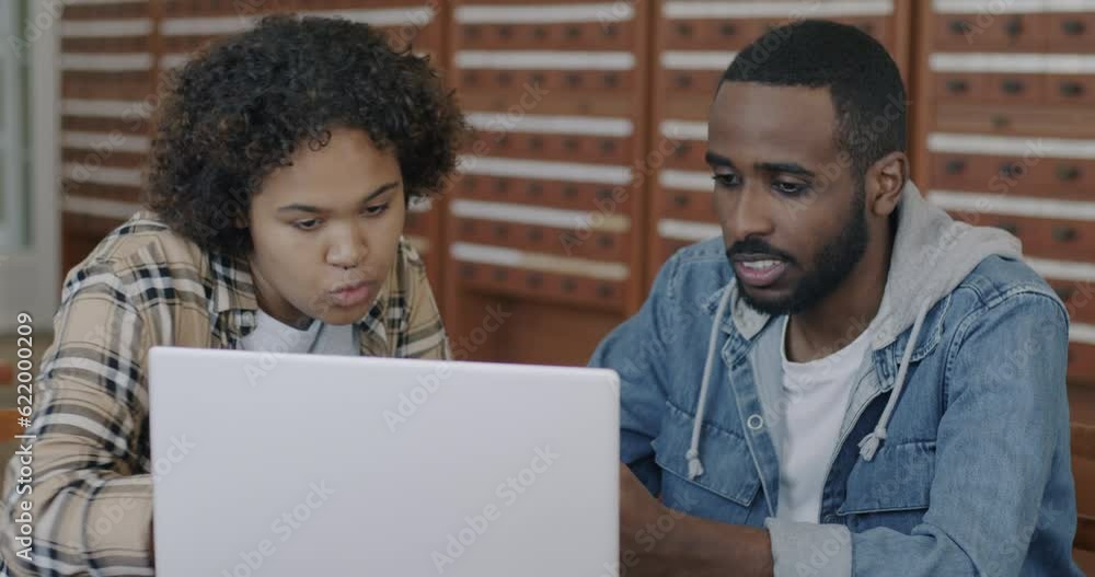 Two African American students man and woman working with laptop computer in library hall talking and looking at screen. Modern technology and education concept. Two African American students man and woman working with laptop computer in library hall talking and looking at screen. Modern technology and education concept.