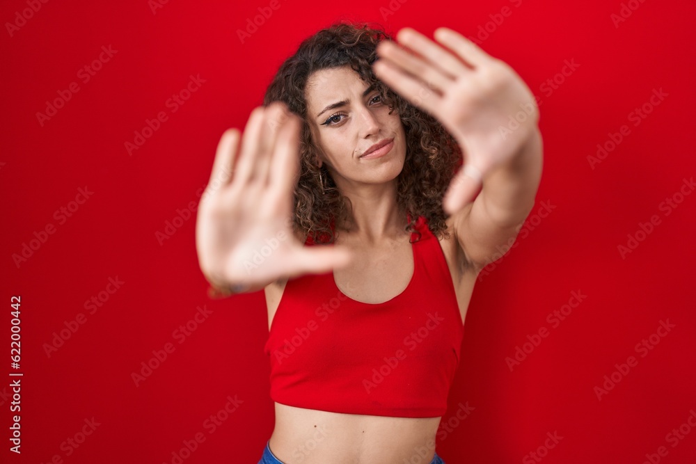 Hispanic woman with curly hair standing over red background doing frame using hands palms and fingers, camera perspective Hispanic woman with curly hair standing over red background doing frame using hands palms and fingers, camera perspective