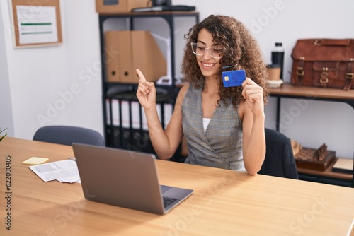 Hispanic woman with curly hair wearing call center agent headset holding credit card smiling happy pointing with hand and finger to the side