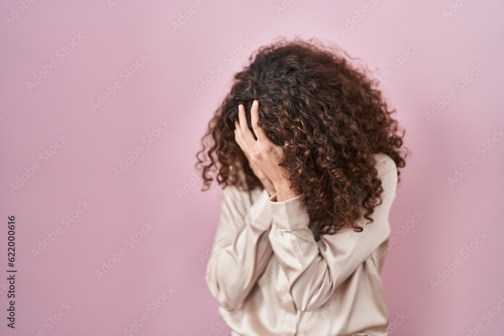 Hispanic woman with curly hair standing over pink background with sad expression covering face with hands while crying. depression concept. Hispanic woman with curly hair standing over pink background with sad expression covering face with hands while crying. depression concept.