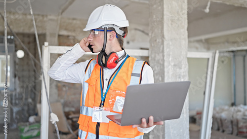 Young caucasian man architect talking on smartphone using laptop at construction site