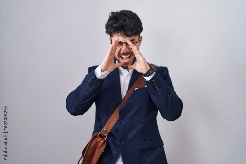 Hispanic man with beard wearing business clothes shouting angry out loud with hands over mouth Hispanic man with beard wearing business clothes shouting angry out loud with hands over mouth