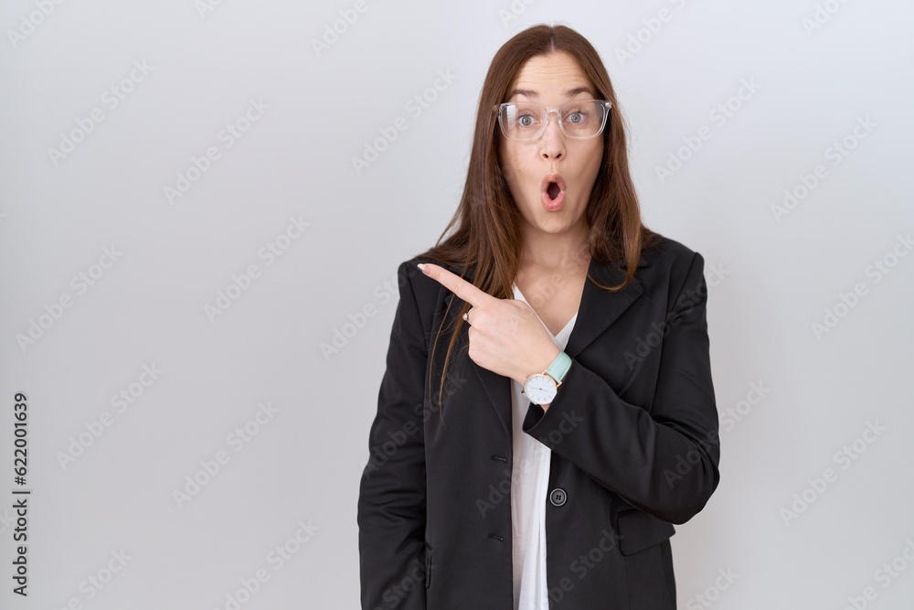 Beautiful brunette woman wearing business jacket and glasses surprised pointing with finger to the side, open mouth amazed expression. Beautiful brunette woman wearing business jacket and glasses surprised pointing with finger to the side, open mouth amazed expression.