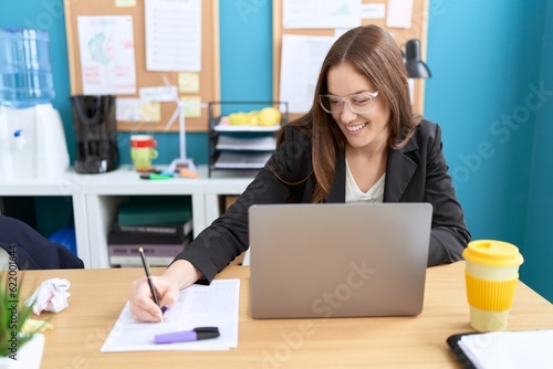 Young beautiful woman business worker using laptop writing on document at office