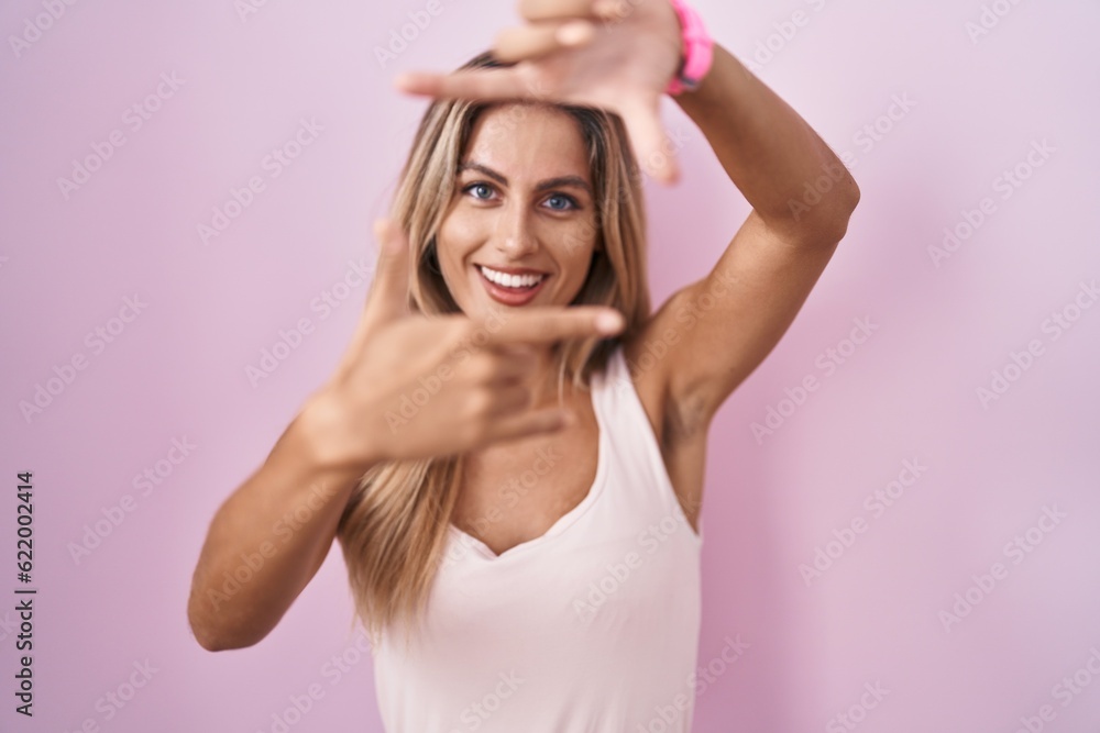 Young blonde woman standing over pink background smiling making frame with hands and fingers with happy face. creativity and photography concept. Young blonde woman standing over pink background smiling making frame with hands and fingers with happy face. creativity and photography concept.