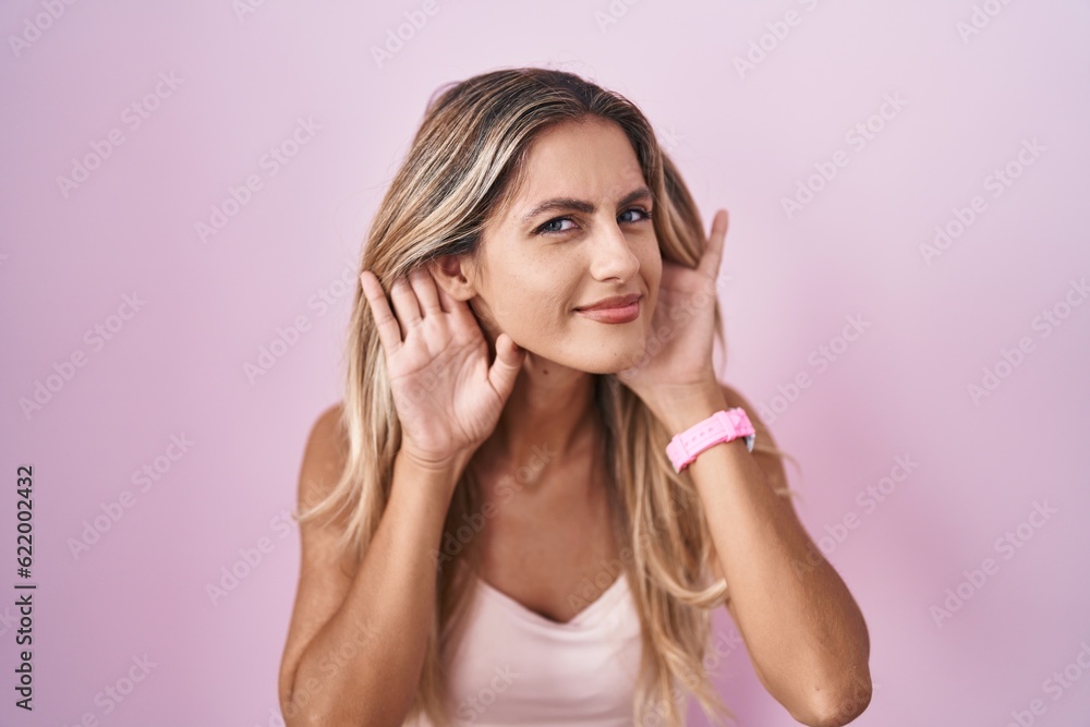 Young blonde woman standing over pink background trying to hear both hands on ear gesture, curious for gossip. hearing problem, deaf Young blonde woman standing over pink background trying to hear both hands on ear gesture, curious for gossip. hearing problem, deaf