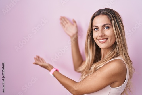 Young blonde woman standing over pink background inviting to enter smiling natural with open hand © Krakenimages.com