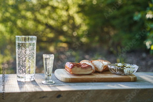 Typical slavic style snack consist from smoked pork bacon, a few slices for rustic bread, pickled sweet and saur cucumbers and shot of vodka and glass of pring water served outside on wooden plate. photo