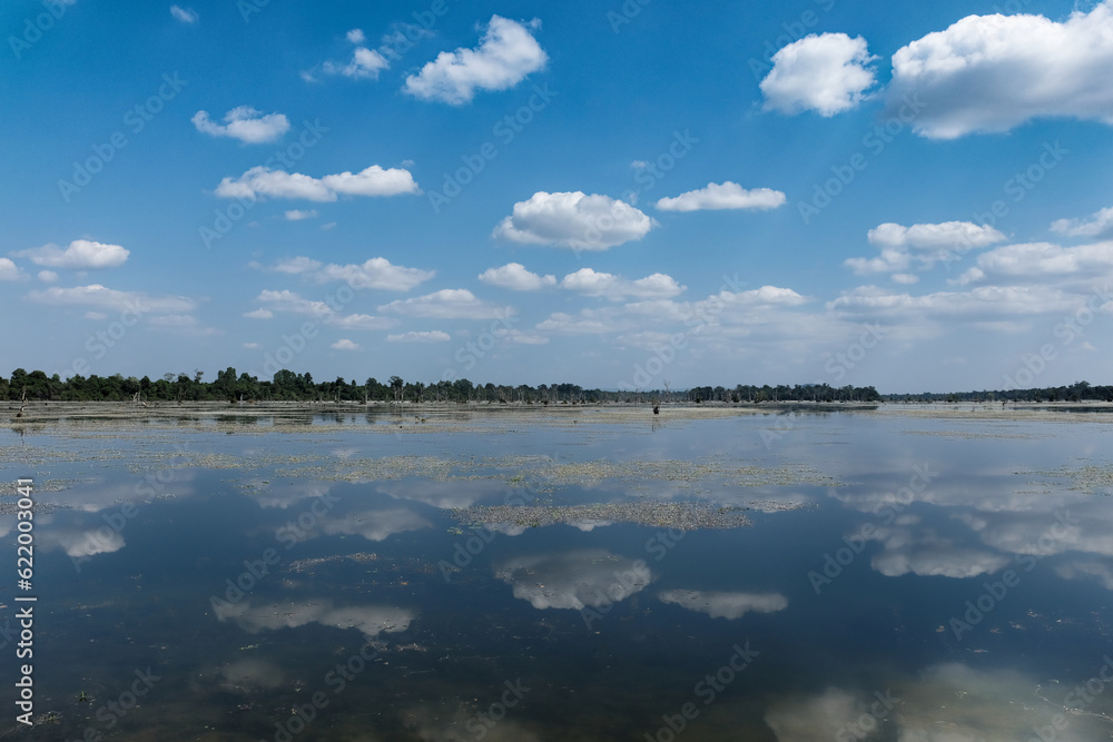 White fluffy clouds are reflected from the calm surface of the Cambodian pond on a sunny day, landscape.