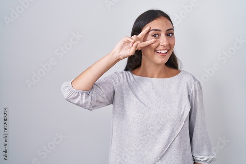 Young hispanic woman standing over white background doing peace symbol with fingers over face, smiling cheerful showing victory © Krakenimages.com