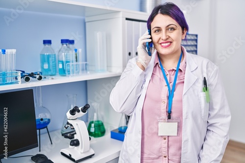 Plus size woman wit purple hair working at scientist laboratory speaking on g the phone looking positive and happy standing and smiling with a confident smile showing teeth