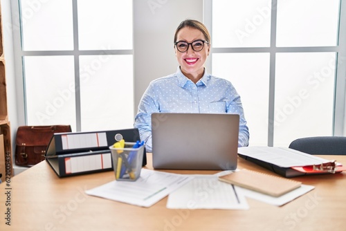 Young hispanic woman working at the office wearing glasses looking positive and happy standing and smiling with a confident smile showing teeth