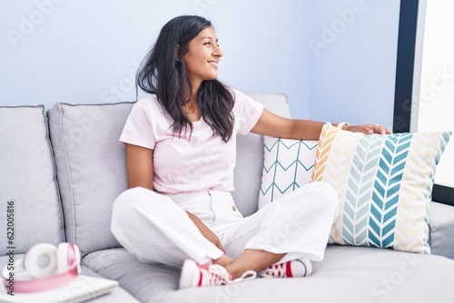Young hispanic woman sitting on the sofa at home looking away to side with smile on face, natural expression. laughing confident.