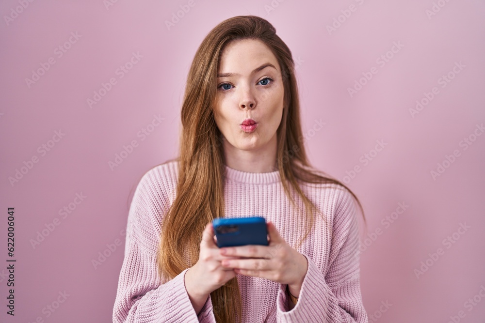 Young caucasian woman using smartphone typing message making fish face with mouth and squinting eyes, crazy and comical. Young caucasian woman using smartphone typing message making fish face with mouth and squinting eyes, crazy and comical.