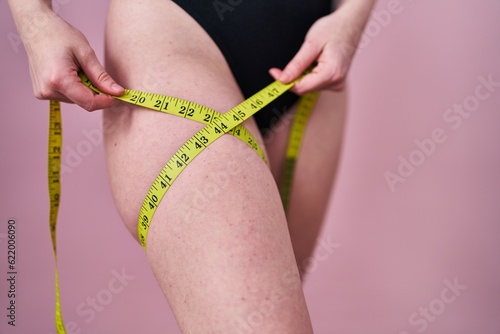 Young caucasian woman measuring leg over isolated pink background