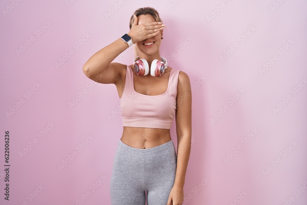 Young blonde woman wearing sportswear and headphones smiling and laughing with hand on face covering eyes for surprise. blind concept.