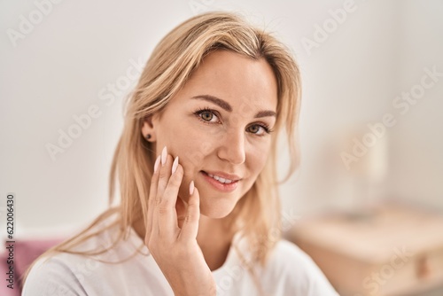 Young blonde woman smiling confident touching face at bedroom