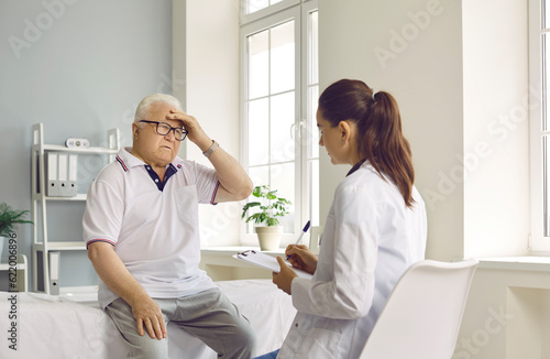 Senior man patient suffering from a headache, sitting on a medical couch, holding hand on his head and telling the doctor about the intense pain that he is feeling. Medicine, medical help concept