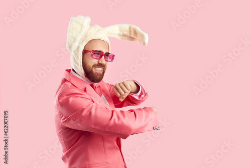 Goofy guy in funny rabbit costume having fun in the studio. Happy confident bearded young man wearing suit, plush Easter bunny ears and pink party glasses dancing isolated on pink colour background