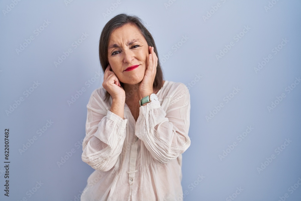 Middle age hispanic woman standing over blue background covering ears with fingers with annoyed expression for the noise of loud music. deaf concept. Middle age hispanic woman standing over blue background covering ears with fingers with annoyed expression for the noise of loud music. deaf concept.