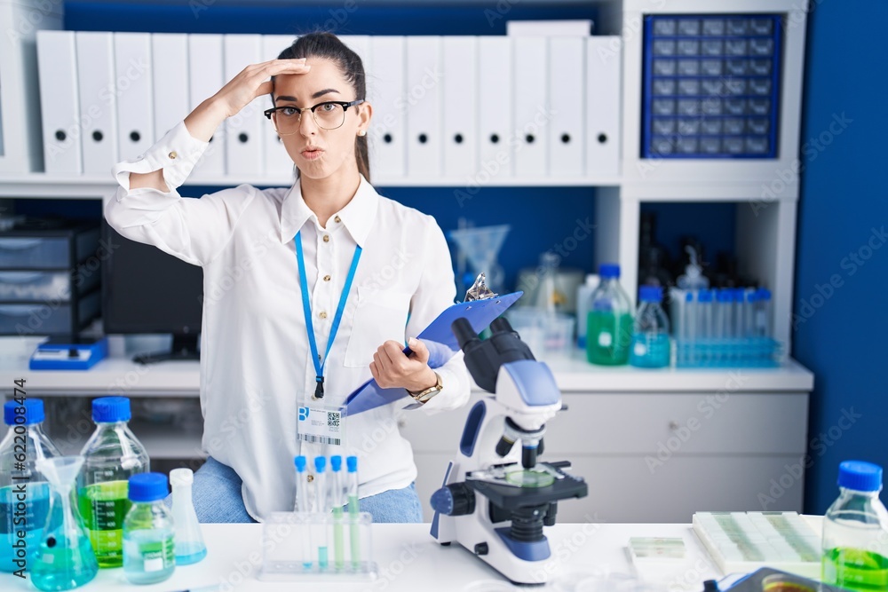 Fototapeta premium Young brunette woman working at scientist laboratory worried and stressed about a problem with hand on forehead, nervous and anxious for crisis