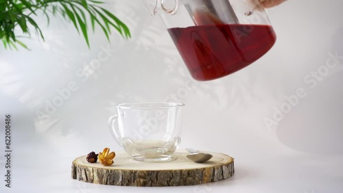 Hot red hibiscus tea is pouring from  teapot into glass cup on white background