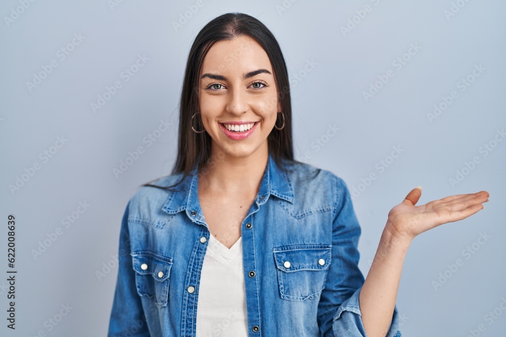 Hispanic woman standing over blue background smiling cheerful presenting and pointing with palm of hand looking at the camera. Hispanic woman standing over blue background smiling cheerful presenting and pointing with palm of hand looking at the camera.