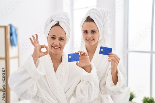 Middle age woman and daughter wearing bathrobe holding credit card doing ok sign with fingers, smiling friendly gesturing excellent symbol