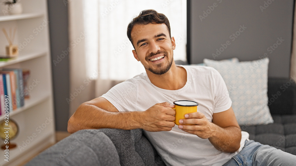 Young hispanic man drinking coffee sitting on sofa at home