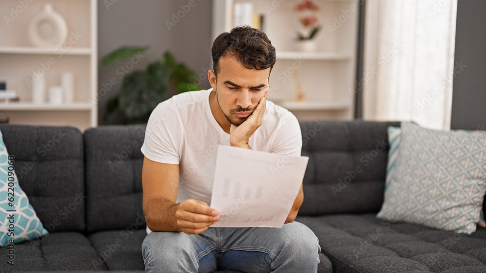 Young hispanic man reading bill sitting on sofa looking upset at home Young hispanic man reading bill sitting on sofa looking upset at home