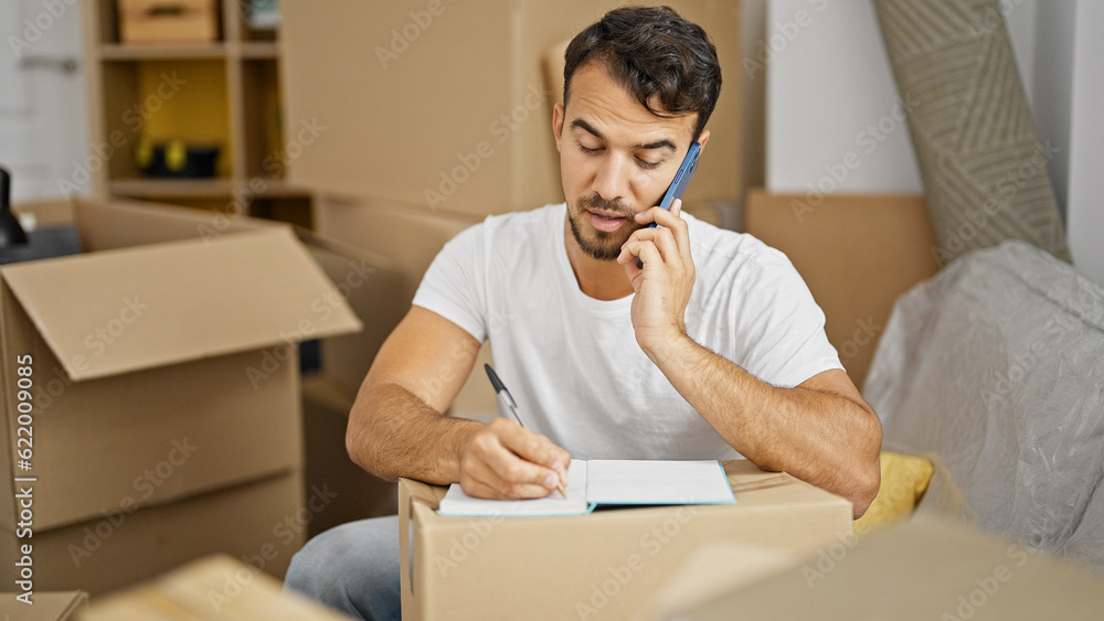 Young hispanic man talking on smartphone taking notes at new home Young hispanic man talking on smartphone taking notes at new home