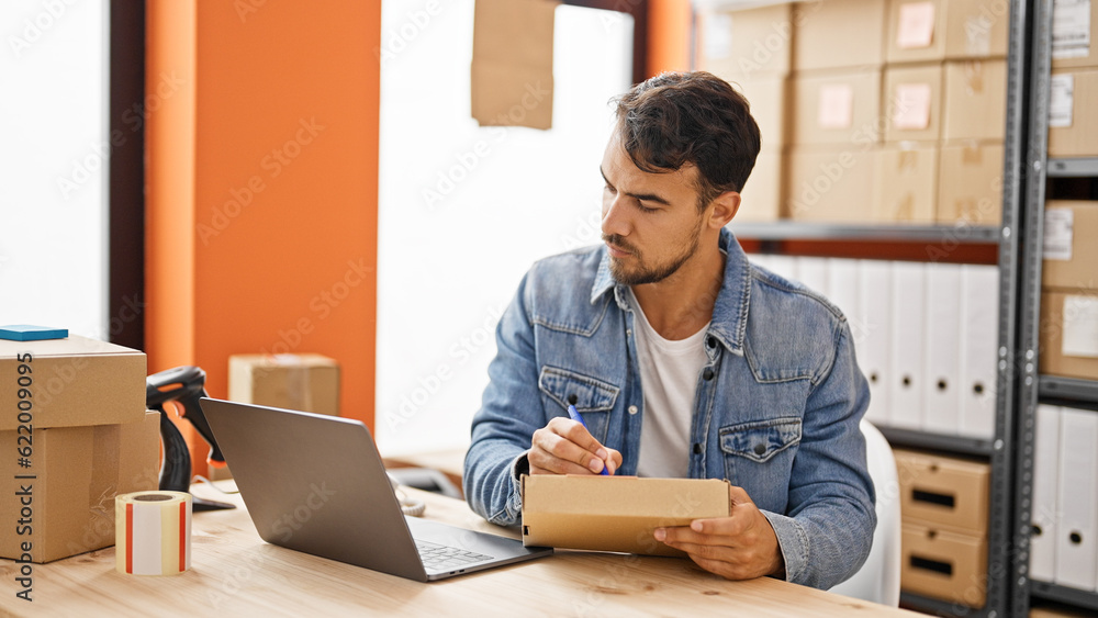 Young hispanic man ecommerce business worker using laptop writing on package at office Young hispanic man ecommerce business worker using laptop writing on package at office