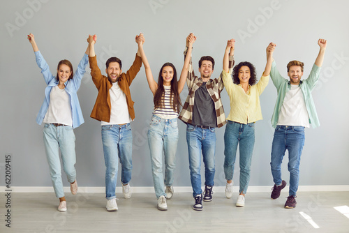 Happy overjoyed friends or coworkers looking at the camera, holding hands up on gray background. Portrait of a group of young smiling people students or colleagues in casual clothes standing together