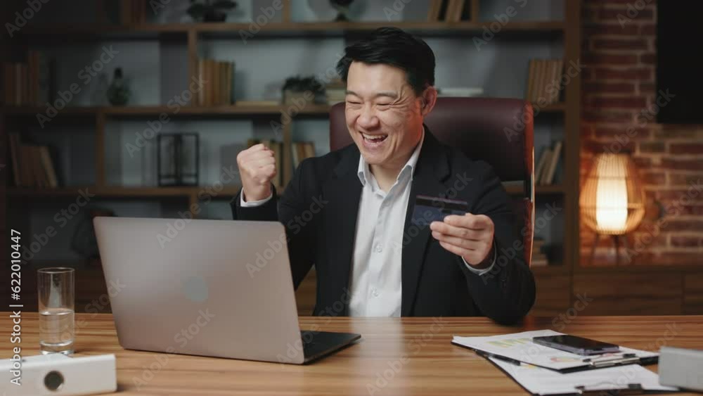 Positive asian employee feeling happy because of successful online payment with credit card on personal computer. Cheerful man making expensive purchase and clenching fist in excitement. Positive asian employee feeling happy because of successful online payment with credit card on personal computer. Cheerful man making expensive purchase and clenching fist in excitement.
